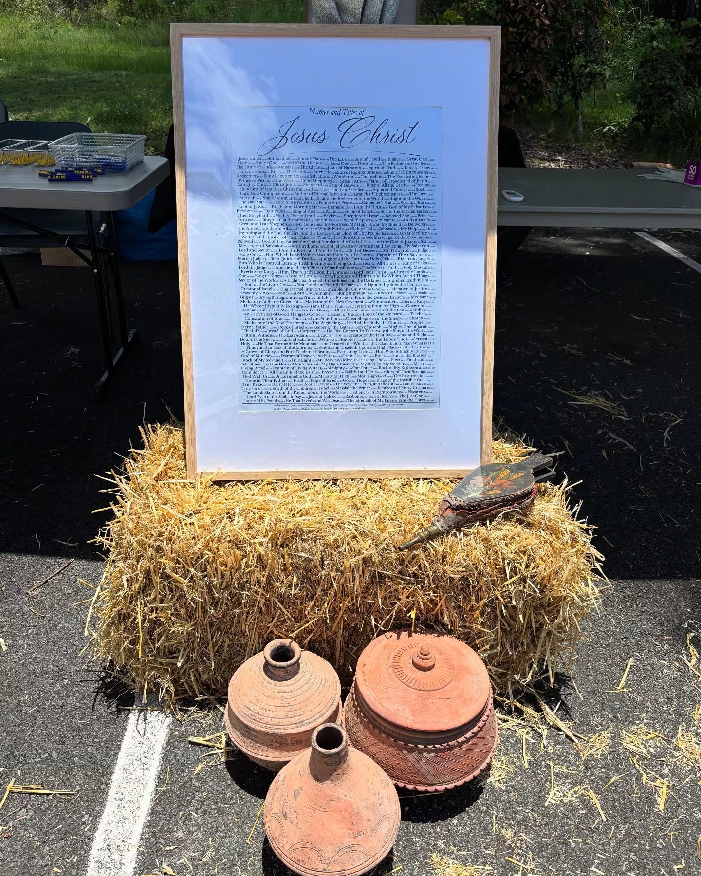 A framed passage about Jesus Christ propped on straw bales beside clay water jars