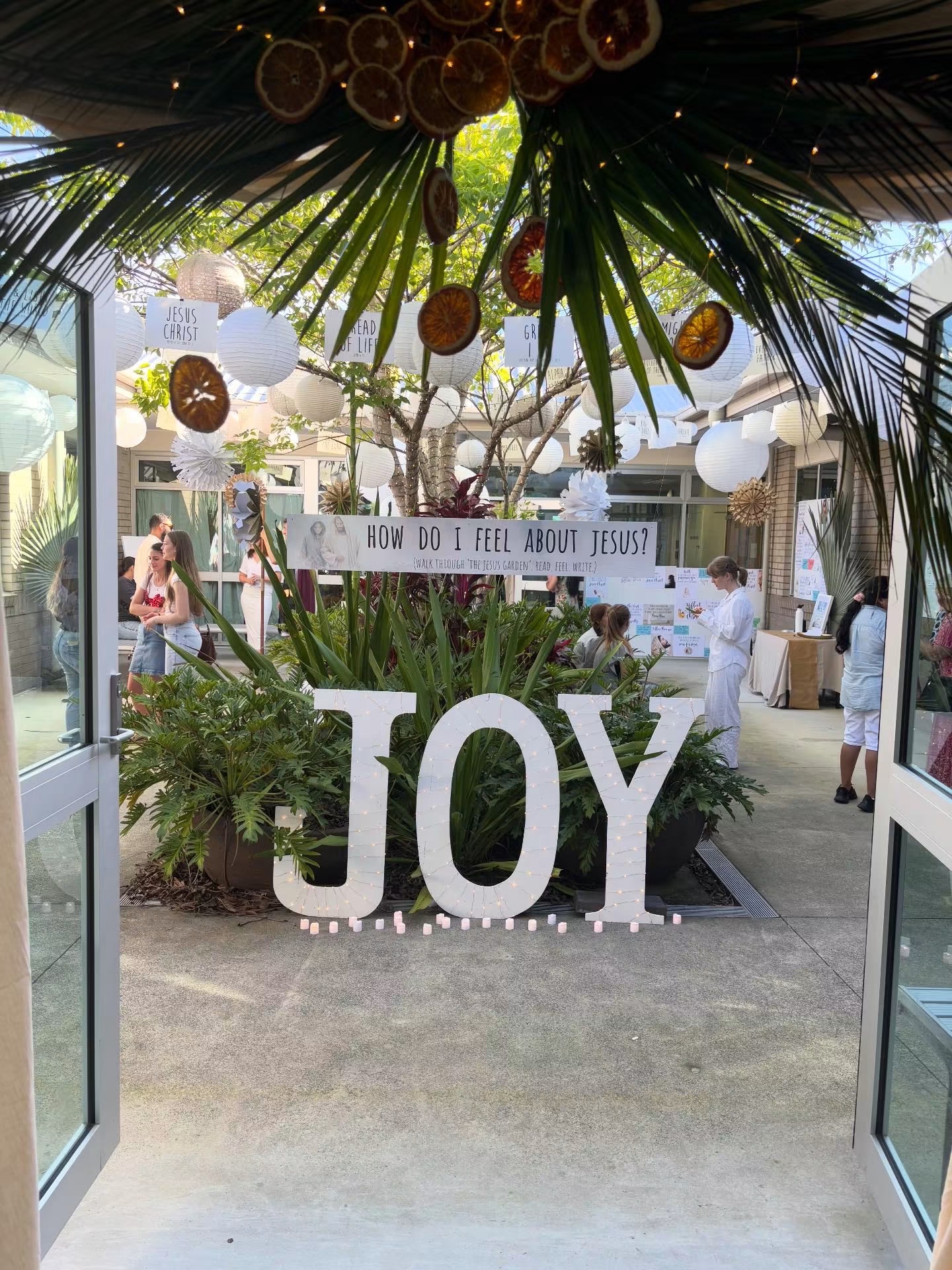The illuminated JOY sign among palms, with paper lanterns overhead